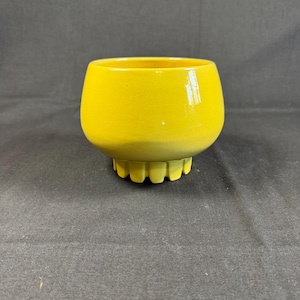 May include: A vibrant yellow ceramic bowl with a rounded shape and a scalloped base. The bowl has a glossy finish and is set against a dark backdrop. This decorative bowl is suitable for holding small items or as a decorative accent.