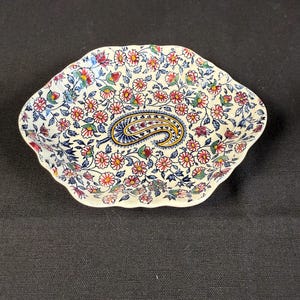 May include: A decorative, floral-patterned dish with a paisley design in the center. The dish features a white background with small pink, red, blue, and yellow flowers. The paisley is blue and gold.