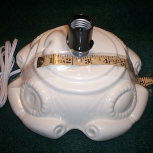 May include: A white ceramic lamp base with a black lamp socket. The base has an ornate design and is approximately 5 inches in diameter. A white electrical cord is attached. A measuring tape is laid across the base.