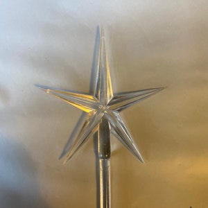 May include: A clear plastic star tree topper with a pointed tip. The star has six points and is designed to be placed on top of a Christmas tree.
