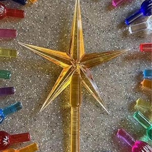 May include: A collection of colorful Christmas light bulbs arranged around a gold star tree topper. The bulbs are in various colors, including orange, blue, purple, green, and pink. The star is a transparent gold color.