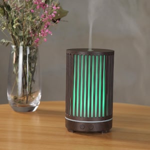 May include: A dark brown essential oil diffuser with vertical light green stripes. The diffuser is cylindrical and has two buttons on the front. A glass vase with pink flowers sits to the left of the diffuser.