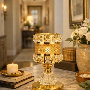 May include: A gold-colored decorative lamp with a crystal base and a cylindrical shade. The shade features a gold mesh center and is adorned with square crystals. A lit candle sits on a small gold tray next to the lamp.