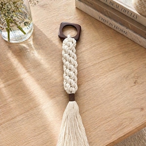 Puede incluir: Un colgante de pared de macramé con una parte superior cuadrada de madera y un diseño de cuerda trenzada de color crema. La cuerda hace la transición a una borla de color crema. El colgante de pared se muestra sobre una superficie de madera marrón claro.
