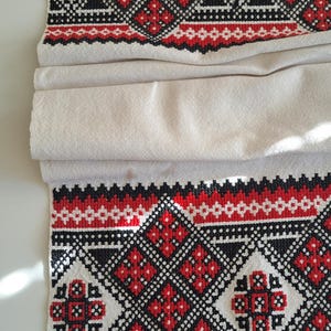 May include: Cream-colored textile with a detailed embroidered pattern in red, black, and white. The design features geometric shapes and floral motifs, creating a traditional folk art aesthetic. The fabric is folded, showcasing the intricate needlework.