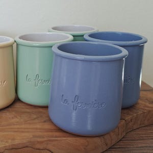 May include: A collection of ceramic jars in various colors, including black, cream, mint green, and blue. Each jar has a rounded shape and the text "la fermière" embossed on the front. They are arranged on a wooden surface.