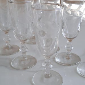May include: A set of six clear glass stemware pieces, each featuring a delicate etched leaf pattern on the bowl. The glasses have a slender cylindrical shape, a short decorative stem with a central knob, and a circular base for stability.