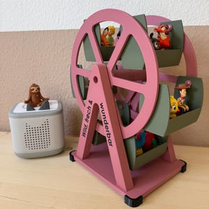 Könnte beinhalten: Ein rosa-grünes Holz-Riesenrad-Spielzeug mit kleinen Charakterfiguren in den Gondeln. Der Text "Wild, frech & Wunderbar" ist auf der Seite aufgedruckt. Links befindet sich ein kleiner Lautsprecher mit einer Faultierfigur.