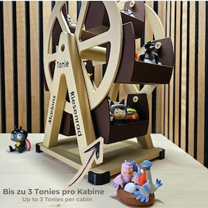 Może przedstawiać: Drewniana zabawka w kształcie diabelskiego młyna z brązowymi i beżowymi akcentami, przeznaczona do małych figurek. Widoczne są napisy "Marlons", "Tonie" i "Riesenrad". Na podstawie zabawki znajduje się napis "Bis zu 3 Tonies pro Kabine".