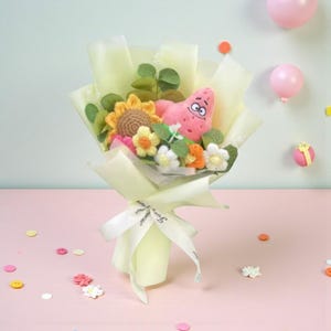 Peut inclure: Un bouquet fantaisiste composé d'une peluche rose en forme d'étoile de mer, d'un tournesol crocheté et de diverses fleurs crochetées colorées. Le bouquet est enveloppé dans du papier vert clair et noué avec un ruban blanc.