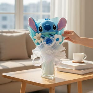 Puede incluir: Un ramo con un personaje de peluche azul de Stitch con orejas grandes, rodeado de flores de tela blancas y azules. El ramo est&aacute; envuelto en papel azul claro y cinta blanca, presentado en un jarr&oacute;n de cristal transparente.
