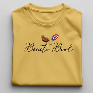 Op de afbeelding: Mosterdgeel T-shirt met de tekst "Benito Bowl" in een sierlijk lettertype. Boven de tekst staan twee kleine afbeeldingen: een geweven mand en een ovaal met het ontwerp van de Puerto Ricaanse vlag.