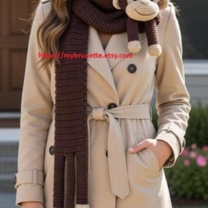 May include: A brown crochet scarf with a monkey design. The scarf is dark brown with long dangling legs and a monkey head with beige accents. The scarf is worn over a tan trench coat.