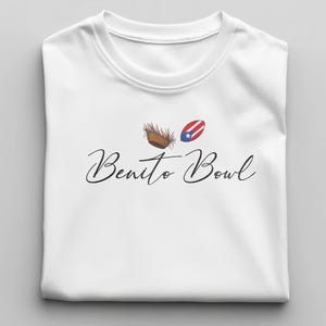 Op de afbeelding: Wit T-shirt met ronde hals en de tekst "Benito Bowl" in een sierlijk lettertype. Boven de tekst staan twee kleine illustraties: een bruin object en een Puerto Ricaanse vlag.