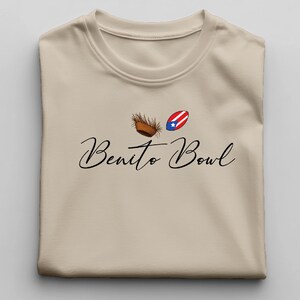 Op de afbeelding: Beige T-shirt met de tekst "Benito Bowl" in een sierlijk lettertype. Boven de tekst staan twee kleine afbeeldingen: een bruine kokosnoot en een Puerto Ricaanse vlag. Het shirt is opgevouwen.