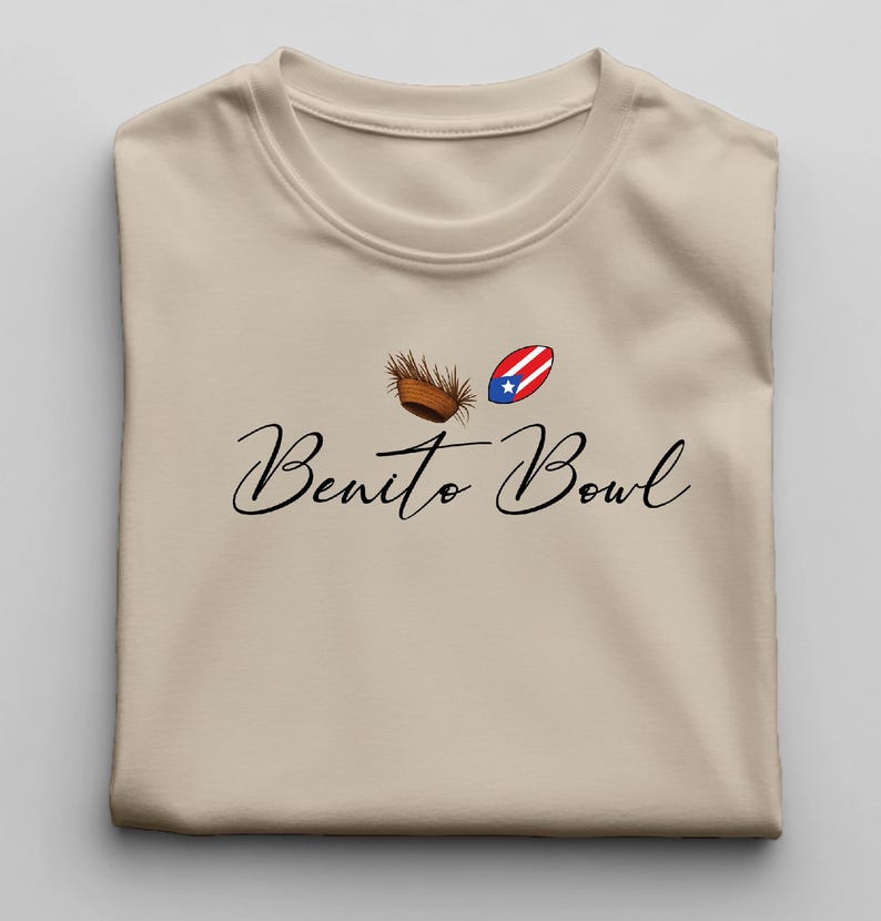 Op de afbeelding: Beige T-shirt met de tekst "Benito Bowl" in een sierlijk lettertype. Boven de tekst staan twee kleine afbeeldingen: een bruine kokosnoot en een Puerto Ricaanse vlag. Het shirt is opgevouwen.
