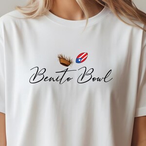 Op de afbeelding: Wit T-shirt met de tekst "Benito Bowl" in cursieve letters. Boven de tekst staan twee kleine afbeeldingen: een bruin, geweven object en een rood, wit en blauw ovaal met een ster. Een casual kledingstuk.