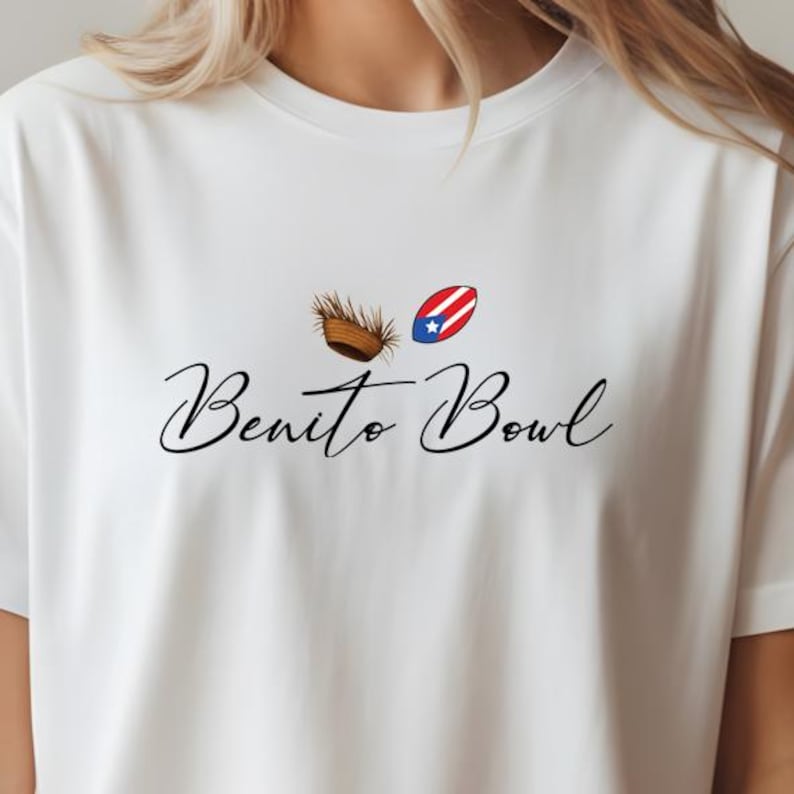 Op de afbeelding: Wit T-shirt met de tekst "Benito Bowl" in cursieve letters. Boven de tekst staan twee kleine afbeeldingen: een bruin, geweven object en een rood, wit en blauw ovaal met een ster. Een casual kledingstuk.