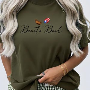 Op de afbeelding: Olijfgroen T-shirt met de tekst "Benito Bowl" in een sierlijk lettertype. Boven de tekst staan twee kleine illustraties: een bruine American football en een American football met het ontwerp van de Puerto Ricaanse vlag.