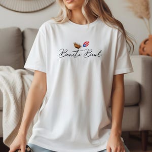 Op de afbeelding: Wit T-shirt met de tekst "Benito Bowl" in een scriptlettertype. Het ontwerp bevat een bruine kom en een rood, wit en blauw grafisch element. Het shirt is een casual stijl met korte mouwen.