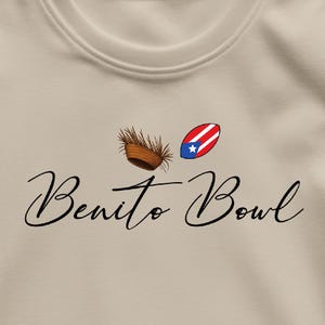 Op de afbeelding: Beige T-shirt met de tekst "Benito Bowl" in cursief. Boven de tekst staan twee illustraties: een geweven mand en een rugbybal met het ontwerp van de Puerto Ricaanse vlag.