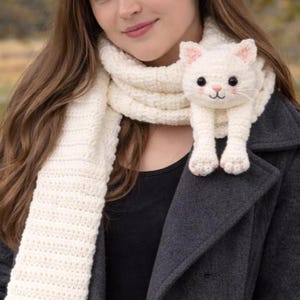 Può includere: Una sciarpa all'uncinetto color crema con una testa di gatto tridimensionale e zampe anteriori attaccate a un'estremità. Il gatto ha occhi a bottone neri, un naso ricamato in rosa e orecchie rosa. La sciarpa ha una trama a coste.