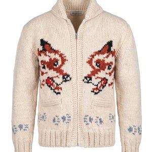 Puede incluir: Un cárdigan de punto grueso color crema con cuello de chal y cremallera frontal completa. El suéter presenta dos motivos simétricos de cabezas de zorro en naranja, negro y blanco, con pequeños patrones de huellas azules cerca del dobladillo.