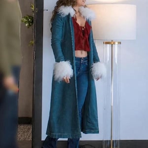 May include: A long teal denim coat featuring a white faux fur collar and white faux fur cuffs on the sleeves. The coat is worn over a dark red ruffled crop top and blue denim jeans. The ensemble is completed with black shoes.