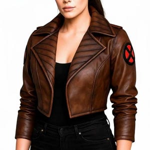May include: A cropped brown leather jacket featuring a wide lapel with horizontal quilted stitching and long sleeves. A circular black patch with a red X-shaped emblem is visible on the left shoulder. The jacket is worn over a black top.