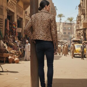 May include: A person wearing a leopard print jacket and black jeans stands on a dusty street. The jacket features a brown and black animal print pattern. The person is wearing black boots. The background shows a street scene with buildings and people.
