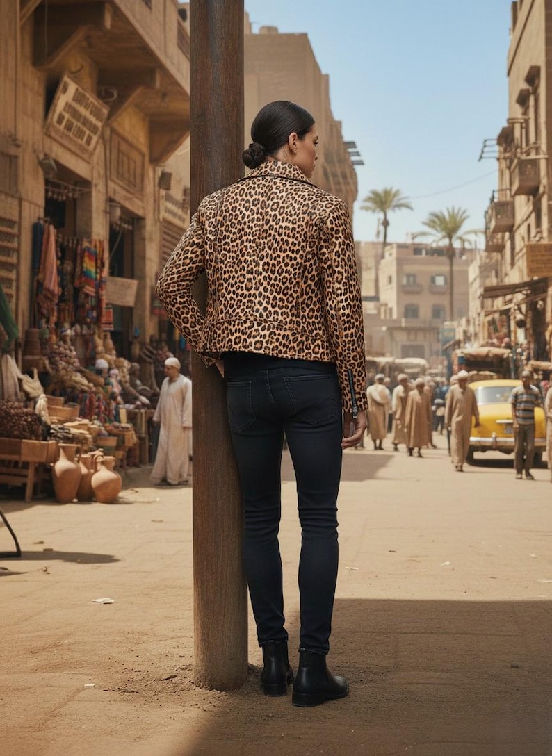 May include: A person wearing a leopard print jacket and black jeans stands on a dusty street. The jacket features a brown and black animal print pattern. The person is wearing black boots. The background shows a street scene with buildings and people.