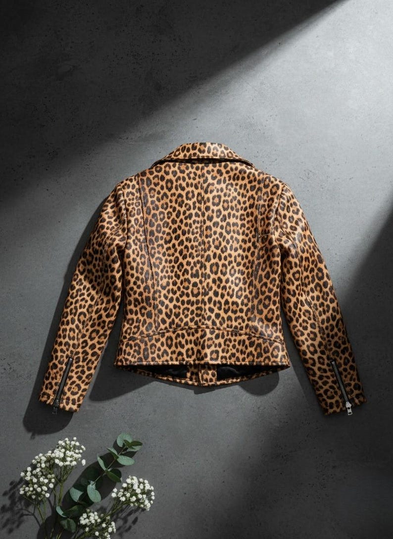 May include: A brown and black leopard print jacket with a classic collar and zipper details on the sleeves. The jacket is made of a faux leather material and has a fitted silhouette. The background is a dark gray.