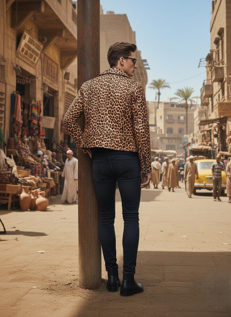 May include: A person wearing a leopard print jacket and dark jeans stands on a sandy street. The jacket features a brown and black spotted pattern. The person is wearing black boots and sunglasses. The background shows a bustling marketplace.