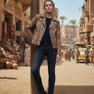 May include: A woman wearing a leopard print jacket, black t-shirt, and dark wash jeans. The jacket has a classic biker style with a notched collar and zipper details. She is standing on a dusty street with a bustling marketplace in the background.
