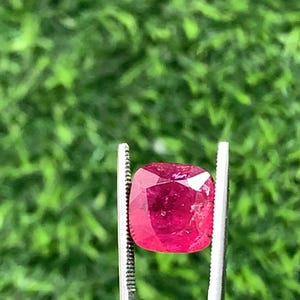 Op de afbeelding: Een kussengeslepen edelsteen met een levendige, dieprode kleur en een gefacetteerd oppervlak, stevig vastgehouden door een zilverkleurige metalen pincet. De steen vertoont interne zuiverheidskenmerken en lichtreflecties op de facetten.