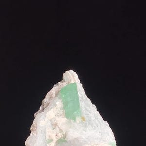 Puede incluir: Una formación de cristal verde claro con forma rectangular se encuentra en una matriz de roca blanca. El cristal es el punto focal, con una apariencia clara y translúcida. La roca tiene una superficie rugosa y texturizada.