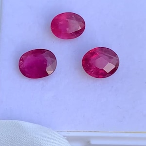 Puede incluir: Tres rubíes ovalados y facetados, de un rojo intenso y vibrante. Las piedras preciosas muestran estructuras internas visibles. Se presentan sobre una superficie blanca, resaltando su brillo. Adecuados para la creación de joyas.