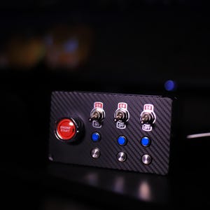 Op de afbeelding: Een zwart bedieningspaneel met een koolstofvezeltextuur. Het heeft een rode "ENGINE START"-knop, drie tuimelschakelaars met de markeringen "ON" en "OFF" en drie blauwe en zilveren knoppen. Ontworpen voor auto- of industrieel gebruik.