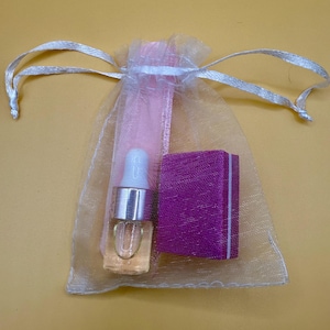 May include: A small glass bottle with a silver cap and a clear liquid, a purple box, and a pink fabric are inside a sheer white drawstring bag. The bag is tied with a white ribbon, all on a yellow background.