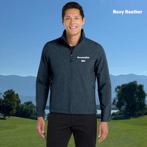 Può includere: Giacca blu navy heather con cerniera intera e collo alto. La giacca presenta la scritta "Personalize Me!" ricamata sul petto. Lo sfondo mostra una persona davanti a un campo verde e un cielo blu.