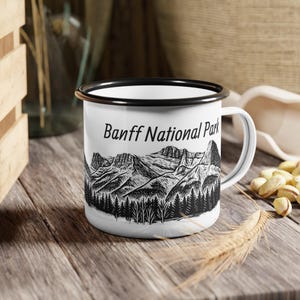 May include: White enamel mug with a black rim and handle. The mug features a black and white illustration of mountains and trees, with the text "Banff National Park" in black. The mug is sitting on a wooden surface.