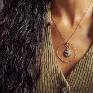 May include: A gold necklace with a circular pendant featuring a gray and pink gemstone. The necklace is worn by a person with long, dark hair and a green sweater.