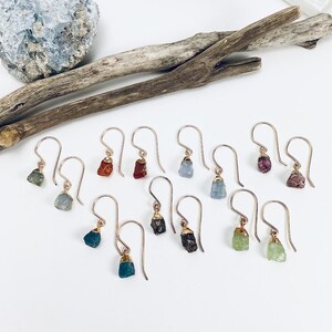 May include: A collection of 10 pairs of earrings featuring different colored gemstones. Each earring has a gold hook and a small, rough-cut gemstone. The gemstones are in shades of blue, green, red, and brown.