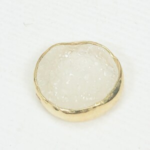 May include: A small, round, white agate stone with a gold-colored metal rim. The stone has a rough, textured surface.