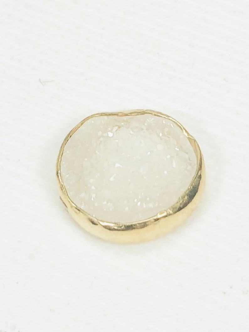 May include: A small, round, white agate stone with a gold-colored metal rim. The stone has a rough, textured surface.