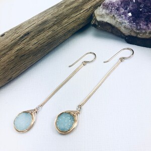 May include: A pair of gold dangle earrings with light blue teardrop-shaped stones. The stones are set in a wire cage and hang from a long, thin gold bar.
