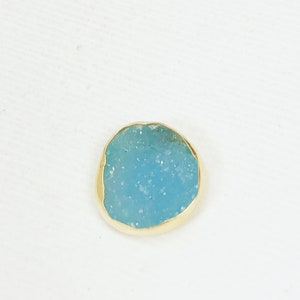 May include: A small, round, light blue stone with a gold rim. The stone has a speckled texture.