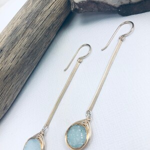 May include: A pair of silver dangle earrings with a light blue gemstone wrapped in gold wire. The earrings are long and slender.