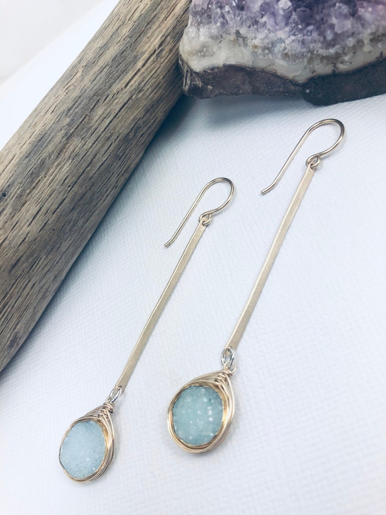 May include: A pair of silver dangle earrings with a light blue gemstone wrapped in gold wire. The earrings are long and slender.