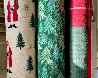 May include: Three rolled-up Christmas-themed fabric cloths. One features Santa Claus and fir trees on a cream background. Another has a green fir tree pattern. The third has a red and green plaid design.